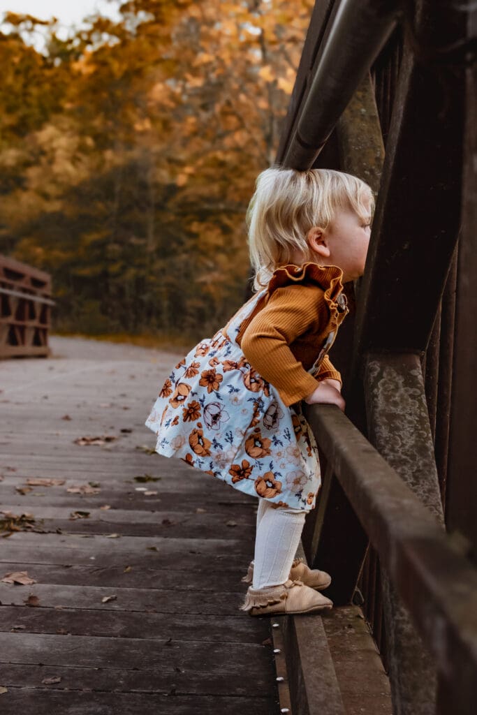 A toddler little girl is standing on a bridge looking through a railing. She's wearing a floral fall outfit with tights. She's in a forested area with fall leaves.