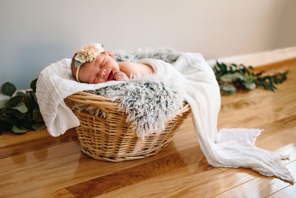 Baby is laying on a white knit blanket on top of a gray fuzzy blanket in a basket. Baby is wearing a floral headband and is smiling while they sleep. In the background there's a gray wall and eucalyptus leaves.