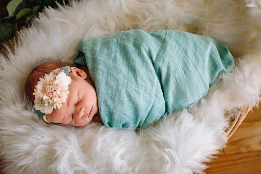 Baby is sleeping peacefully while wearing a floral headband and wrapped in a teal blanket. They're laying on a very fluffy white blanket.