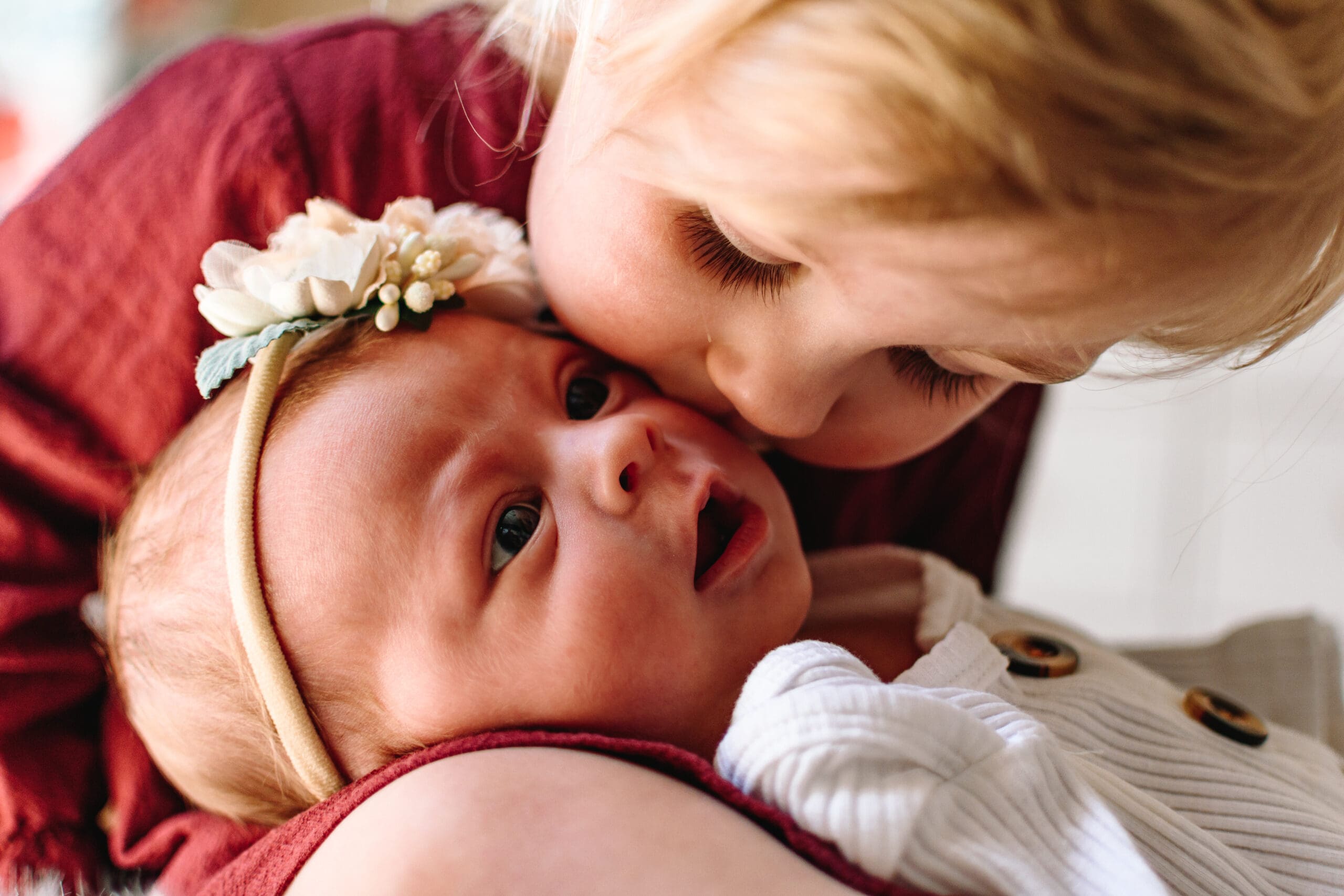 A newborn baby is laying in the lap of a toddler. The toddler is wearing a red dress and baby is wearing a floral headband and a white outfit. Baby is looking up while toddler is kissing their cheek.