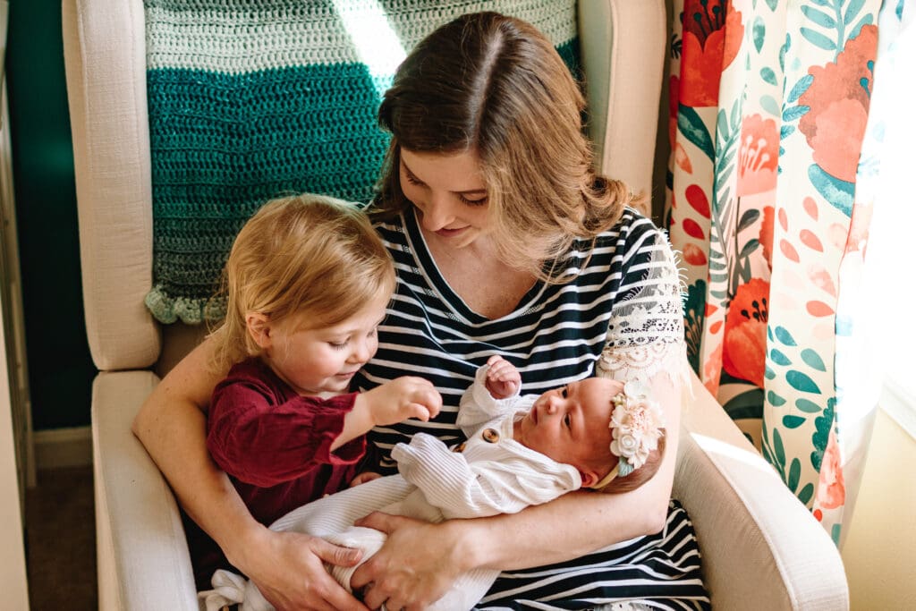 Mom is sitting on a white chair with a green blanket on the back and bright poppy loral curtains in the background. Mom is holding a newborn baby in one arm and has other arm around an older toddler and smiling down at them. toddler is interacting with baby and smiling at her.