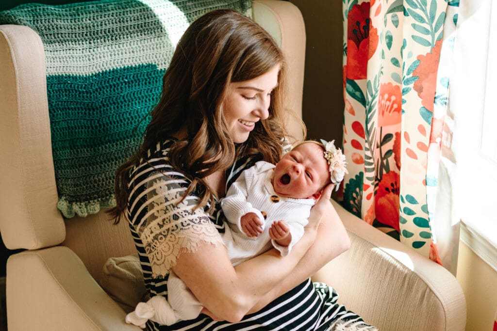 Mom is sitting on a white chair with a green blanket on the back and bright poppy loral curtains in the background. Mom is holding baby as baby yawns and is smiling down at baby.