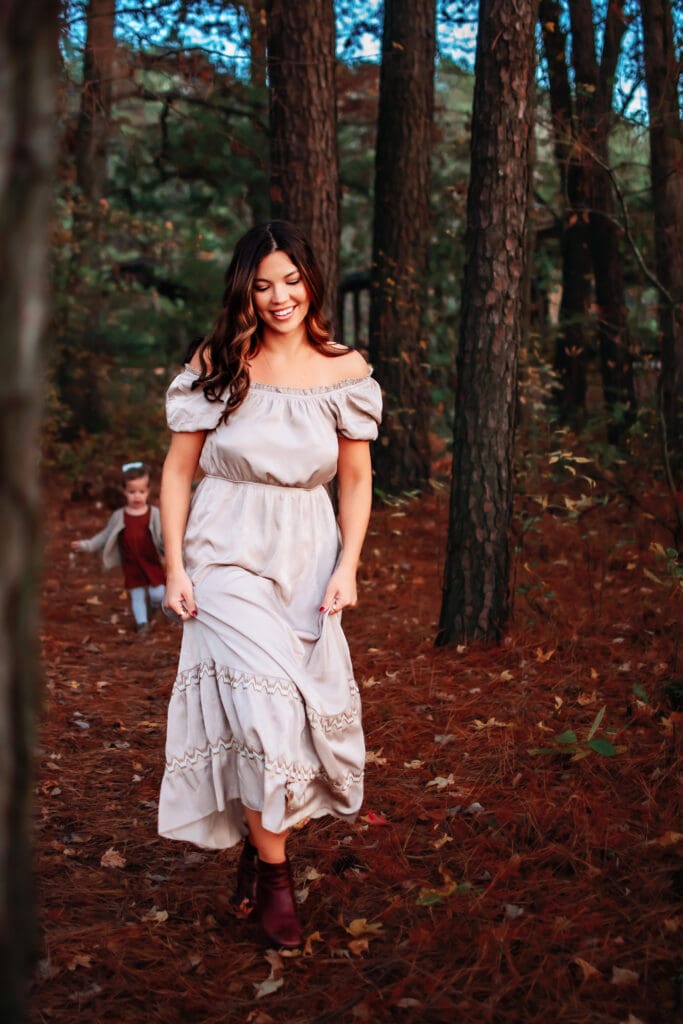 Mom and toddler are in a forested area with trees and leaves on the ground. Mom is in the front walking toward the camera in a long flowy tan dress. Toddler is running behind her in the distance.