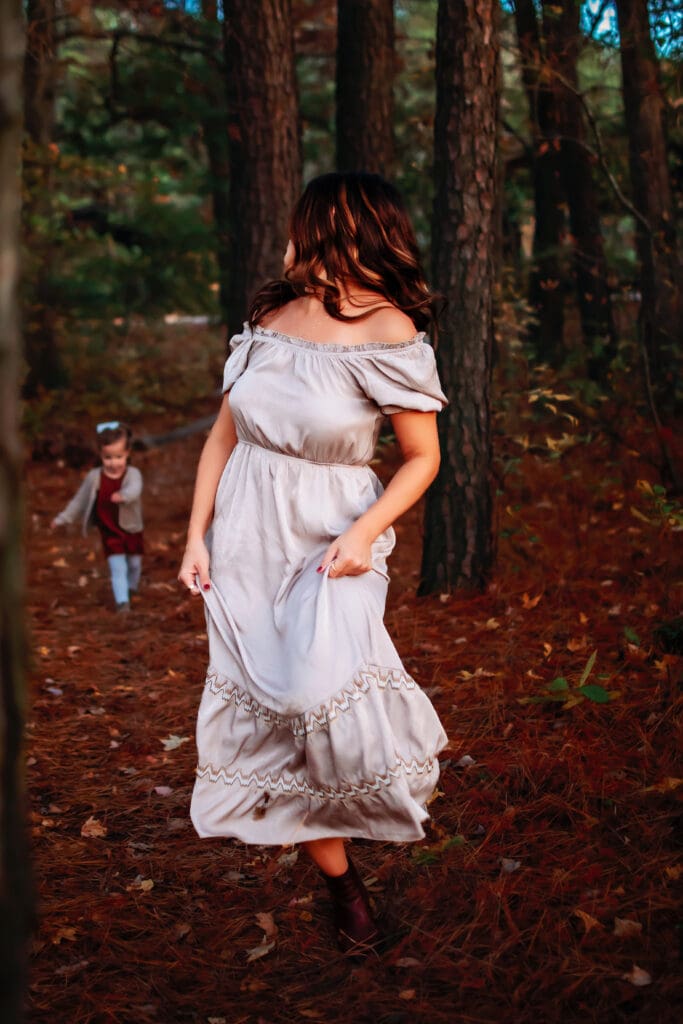 Mom and toddler are in a forested area with trees and leaves on the ground. Mom is in the front walking toward the camera in a long flowy tan dress. Toddler is running behind her in the distance.