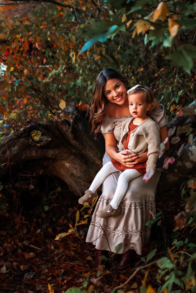 Mom is smiling at the camera wearing a tan flowy long dress holding a toddler in a dress and cardigan and tights. They're sitting on a large log with bright colorful fall trees in the background. 