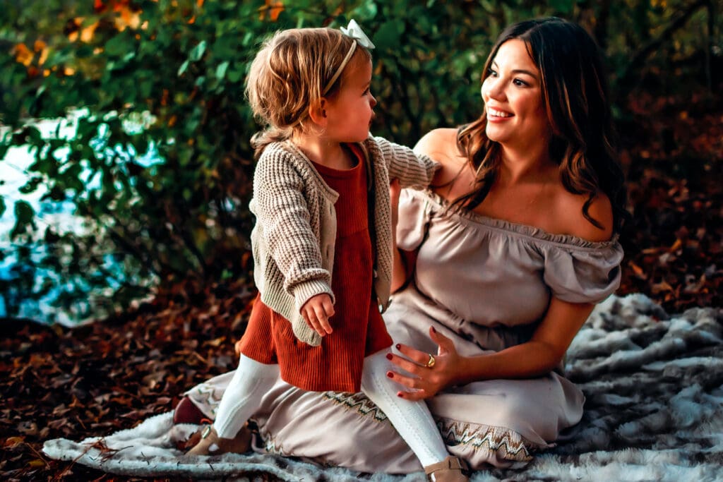 Mom is wearing a long flowy tan dress sitting on a blanket with a fall leaves behind her. There's a toddler in front of her talking to her wearing a burnt orange dress, cardigan, and tights. 