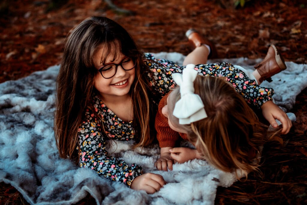 Two little girls are laying on a blanket while looking at each other and giggling.