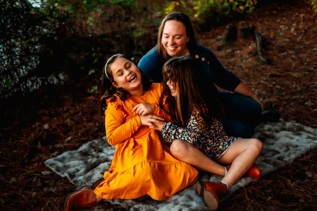 Two little girls are tickling each other and belly laughing. Their mom is sitting behind them smiling at them. They're all sitting on a furry blanket in a wooded area.