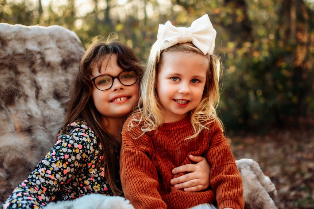 Two little girls are sitting in a furry chair smiling at the camera for a fall family photoshoot.