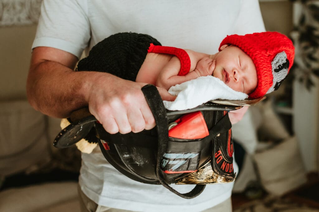 Baby is dressed in a little knit fighter fighter outfit and is laying on a blanket on a firefighter helmet. Babies hand is touching his face and he appears to be sleeping. Dad is dressed in a white shirt and is holding the helmet.