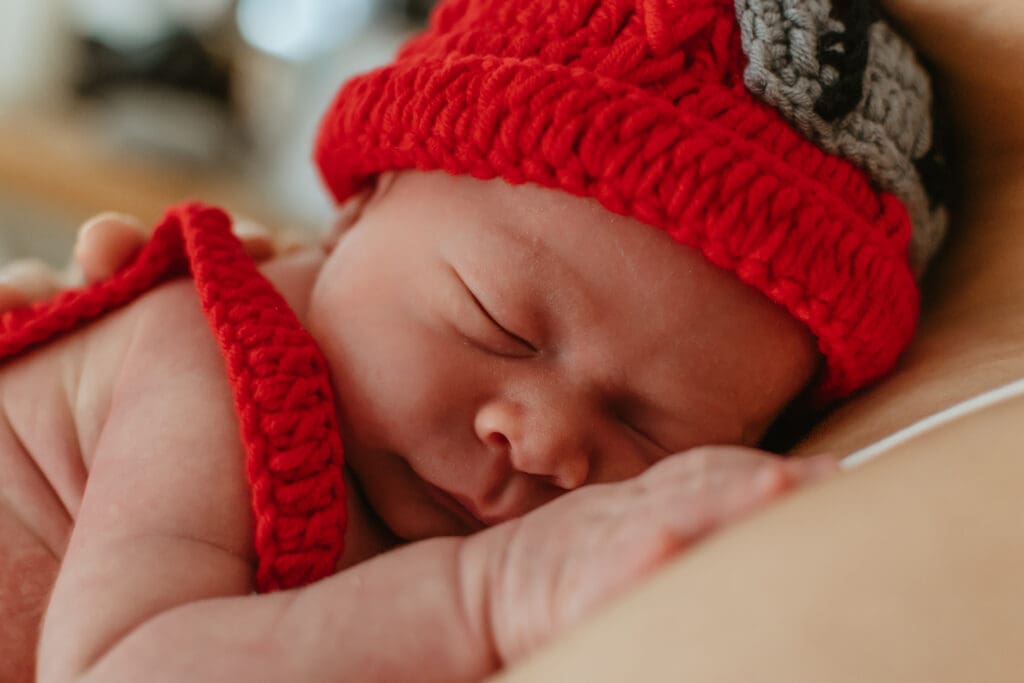 Close up shot of baby laying against moms chest. He's wearing a red firefighter knit had.