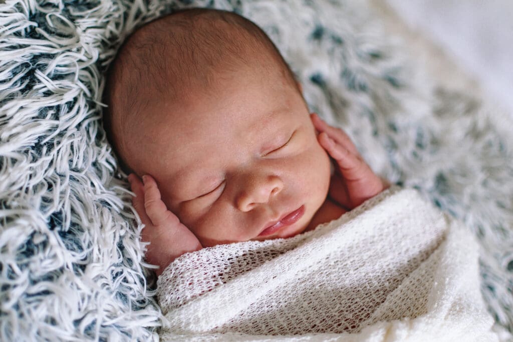 Baby is laying on a fuzzy blanket and appear sleeping while his hands touch his face. He's wrapped in a white blanket.