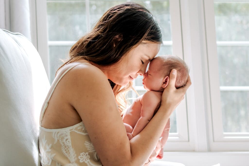 Mom is sitting on a chair in front of a window. She has her eyes closed and is touching foreheads cuddling her newborn baby.