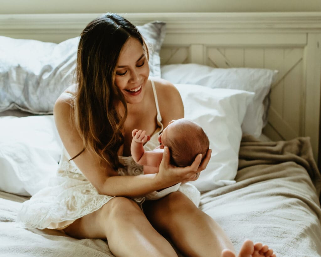 Mom is sitting on a bed with pillows and headboard behind her. She's smiling while looking at baby in her arms facing her.