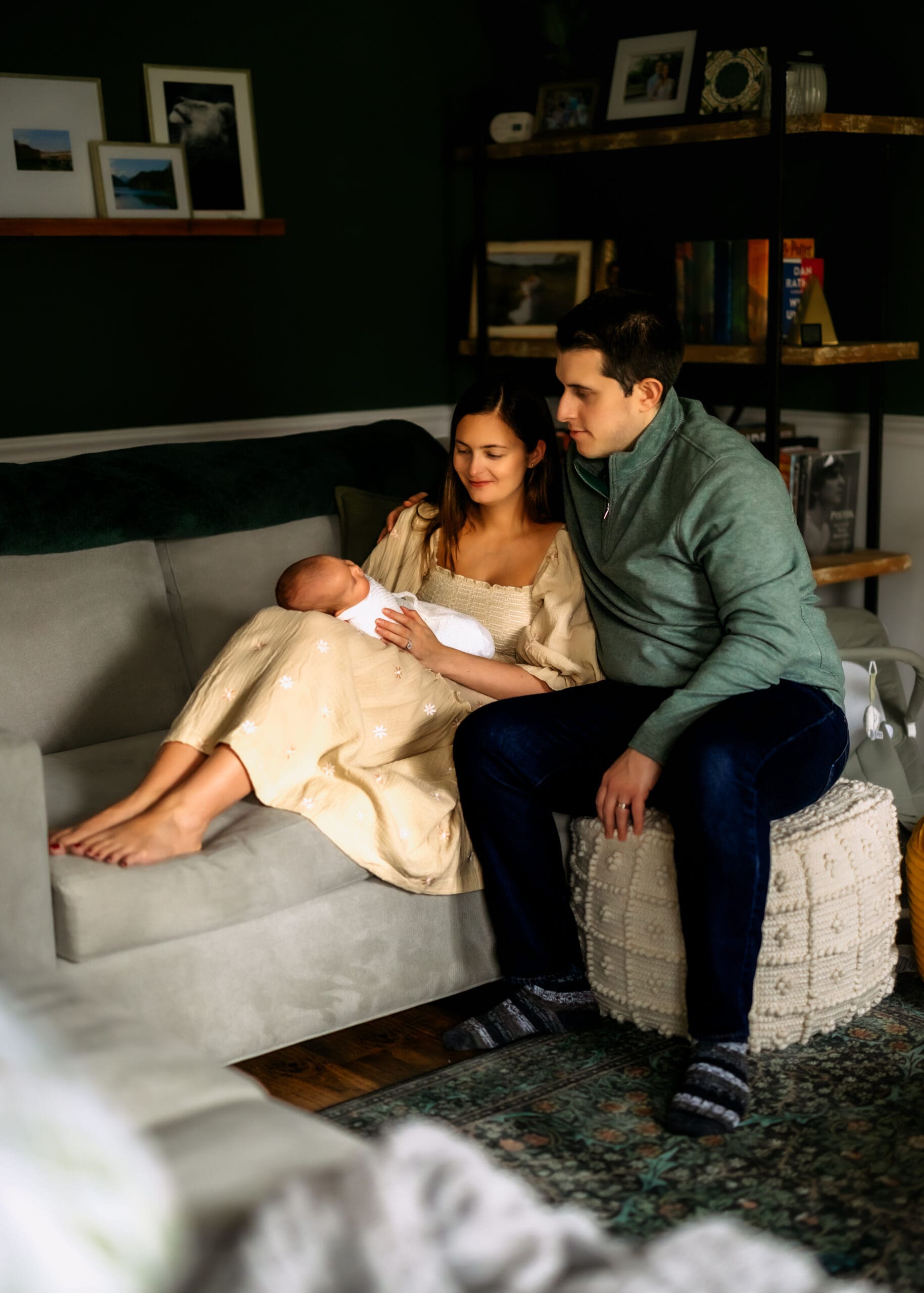 Mom and dad sit in the living room on the couch and ottoman. Moms legs are up on the couch and baby is resting on the tops of her legs. Mom and dad are looking down at baby sweetly.