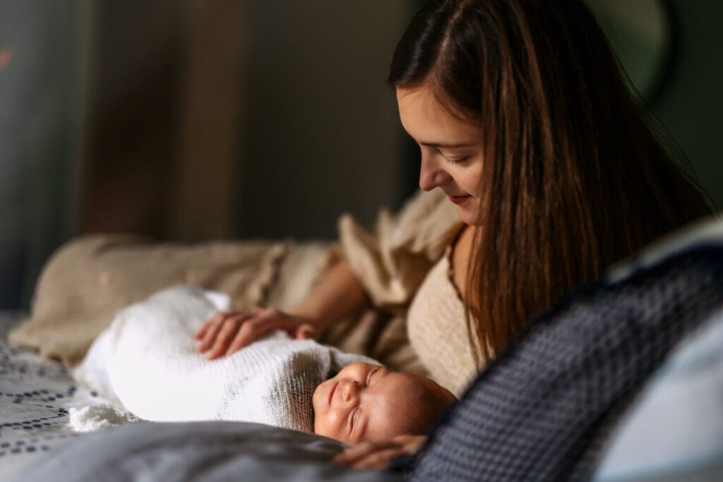 A mom with long hair is sitting on a bed, looking down at a baby wrapped in a white blanket. The baby is lying on the bed, appearing to be asleep with a slight smile on the face. The background is blurred, focusing attention on the mom and baby in this in home newborn photography session.