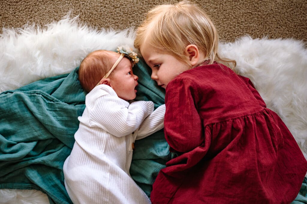 A newborn and an older toddler lay on top of a green blanket on top of a white blanket on top of tan carpet. The baby is wearing a white outfit and headband and the toddler is wearing a red dress. They're cuddled close together looking at each other.