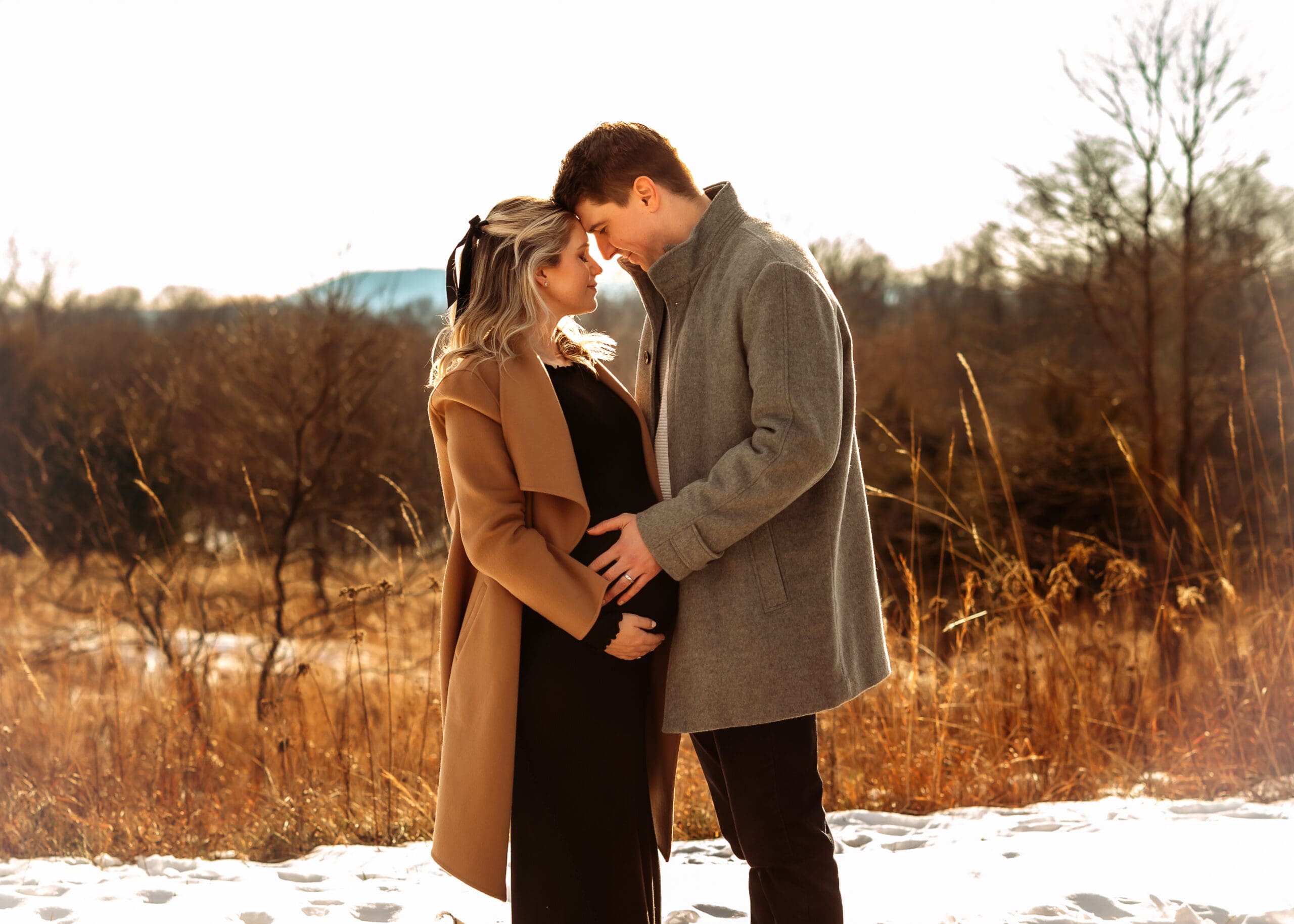 A woman and man stand with their foreheads touching. The woman is pregnant with a growing belly and all hands are touching the stomach. They're standing in a field with tall grass, trees, and mountains in the back.